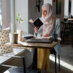 A young woman in a hijab reading a book at a cozy cafe table by the window.